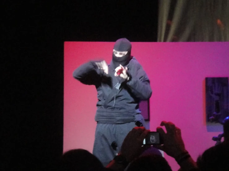 a masked man in front of a crowd is shown with his head down