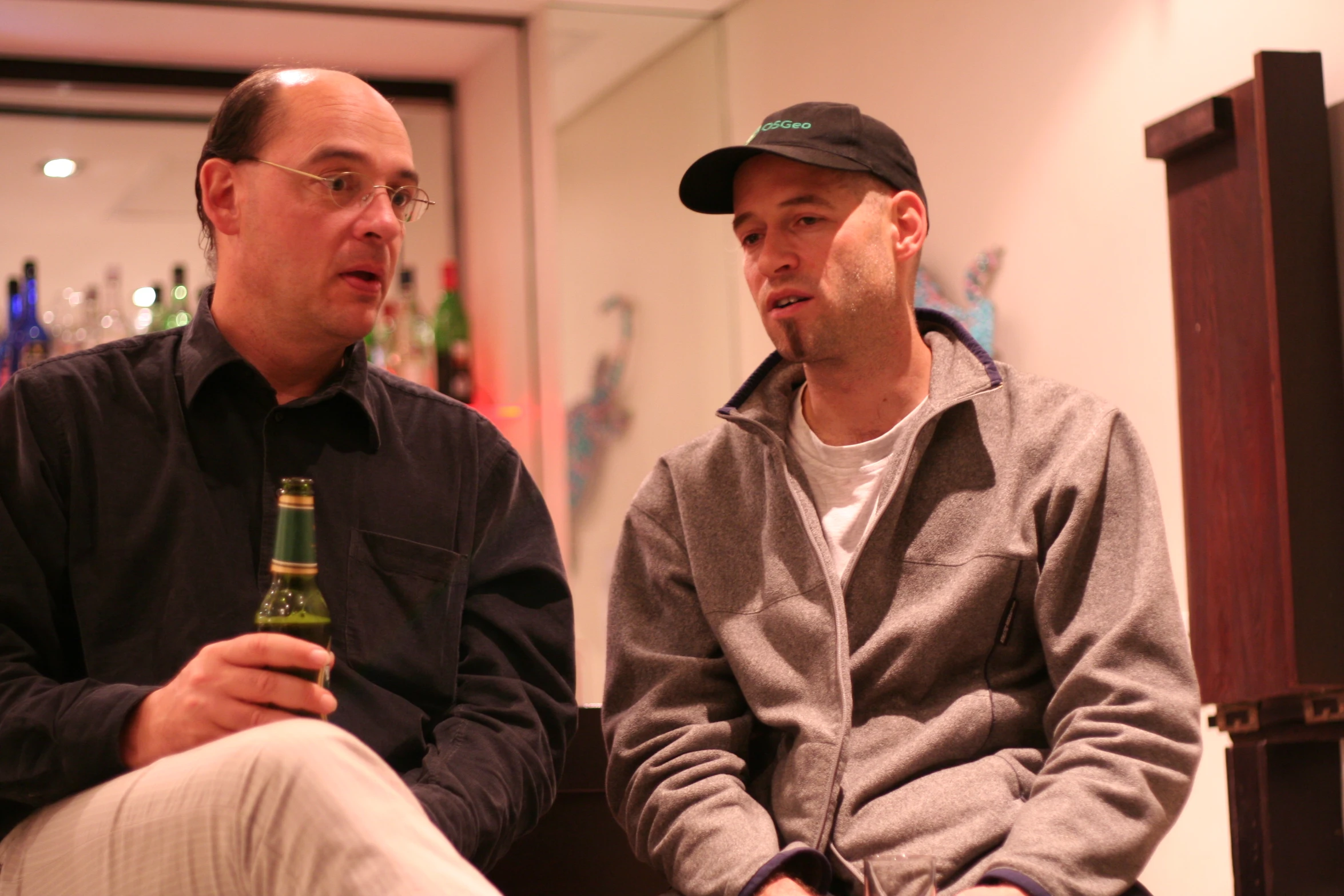 two men sitting down next to each other holding beers