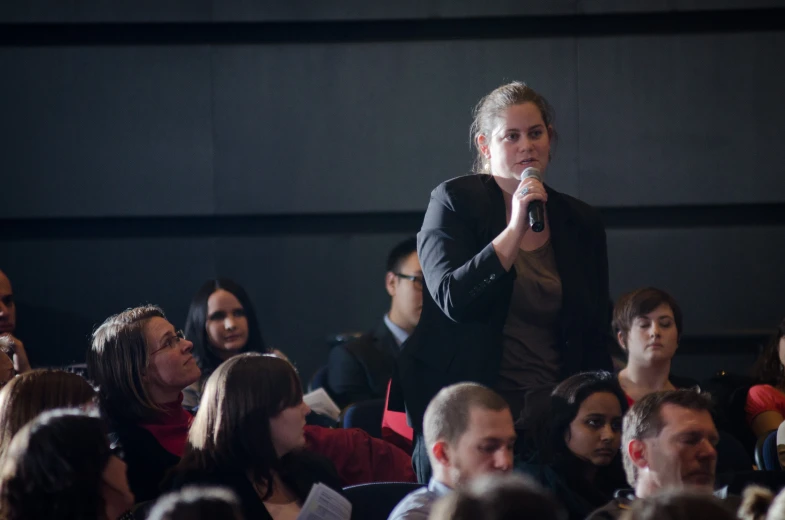 a woman that is talking into a microphone in a room with other people