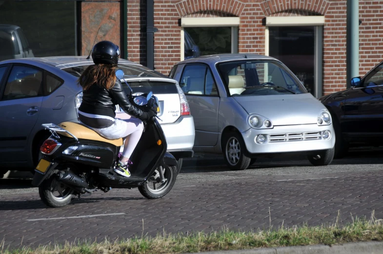 a woman in black jacket riding a scooter on street next to parking lot