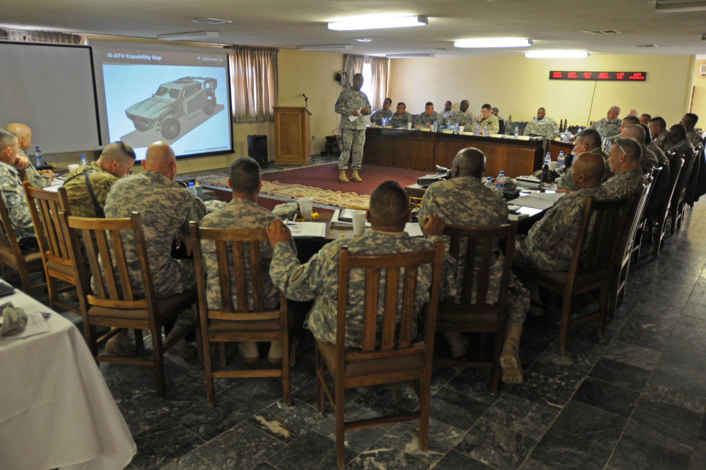 military personnel sit at tables with a presentation in the background