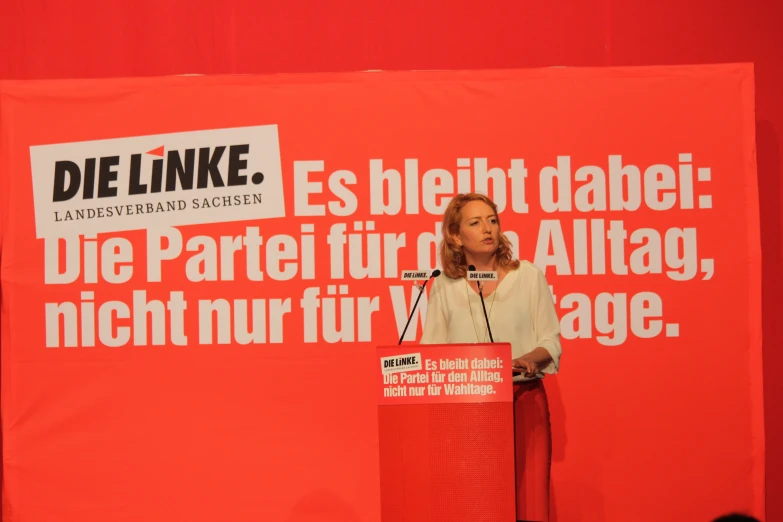 a woman stands at a podium in front of a banner that reads die link
