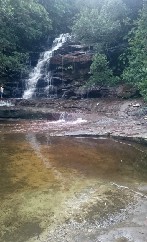 a man stands at the edge of a small lake with a waterfall in the background