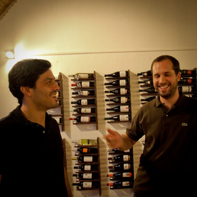 two men standing next to a wall with wine bottles on it