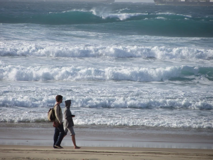 two people walking down the beach near some rough ocean waves