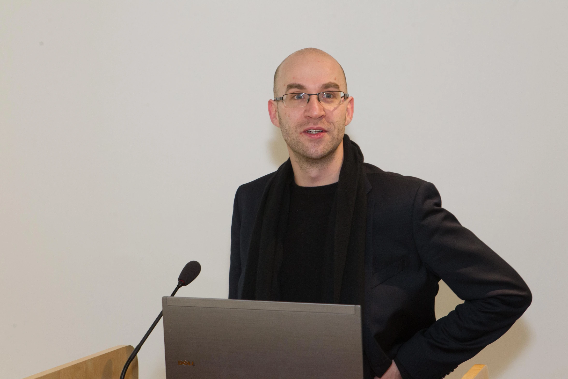 a bald man with glasses speaks into a microphone and holds a laptop