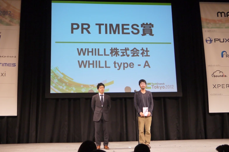 two men standing on a stage with a large screen in front of them