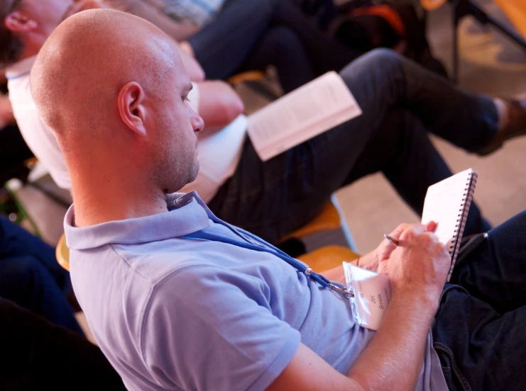 the man in blue shirt is taking notes while another man sits on chair