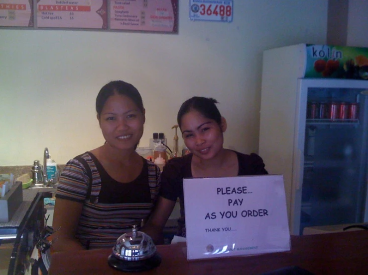 two women standing at the counter in front of a sign that says please pay as you order