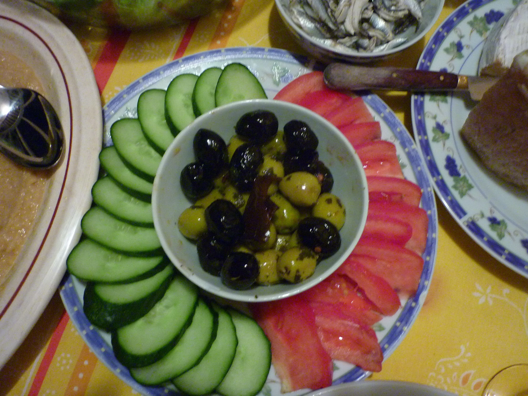 a bowl full of olives, cucumber and tomatoes with salad in the background