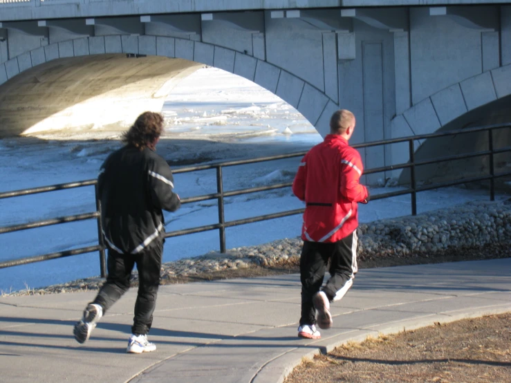 two men run up the sidewalk toward the water under a bridge