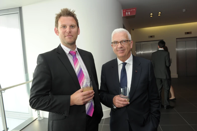 two men in suits stand side by side with a drink