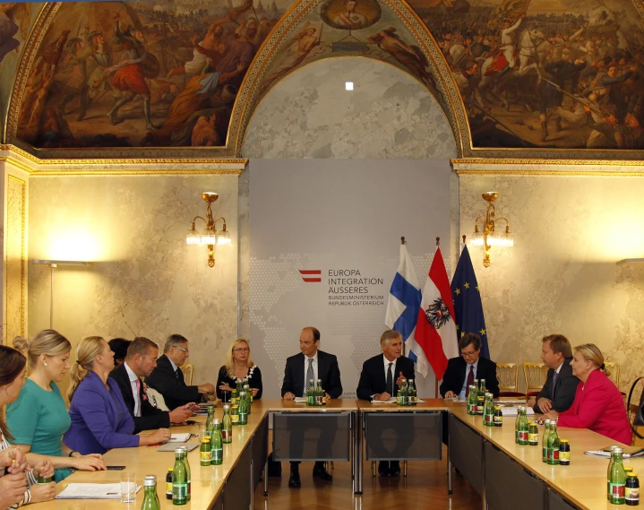 several people in business attire sit around a table while a flag is seen above