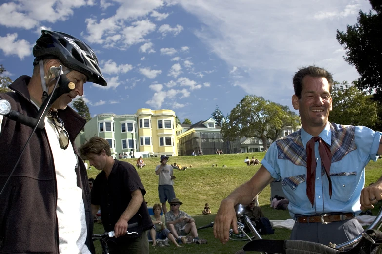 two men wearing helmets standing in the grass while one of them is on his bicycle