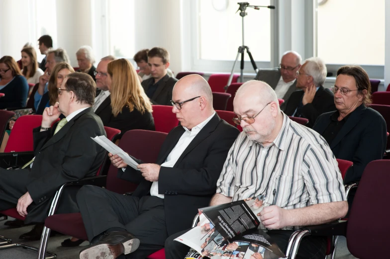 an older man reads a magazine in front of a meeting room full of people