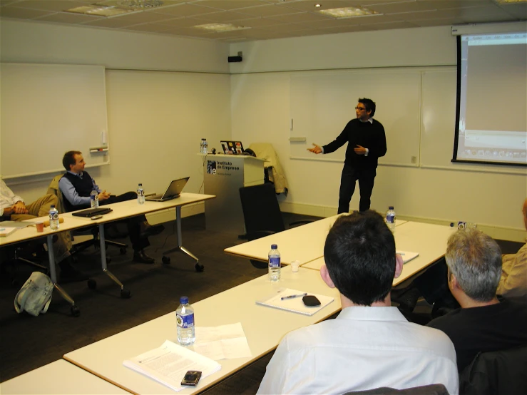 a man stands in front of several people sitting at desks with a projector screen on the wall