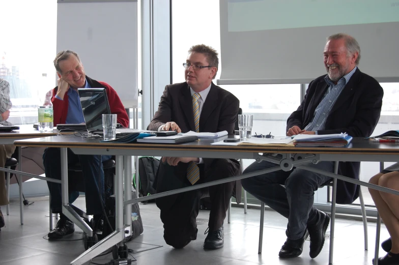 three men are seated at a table discussing a topic