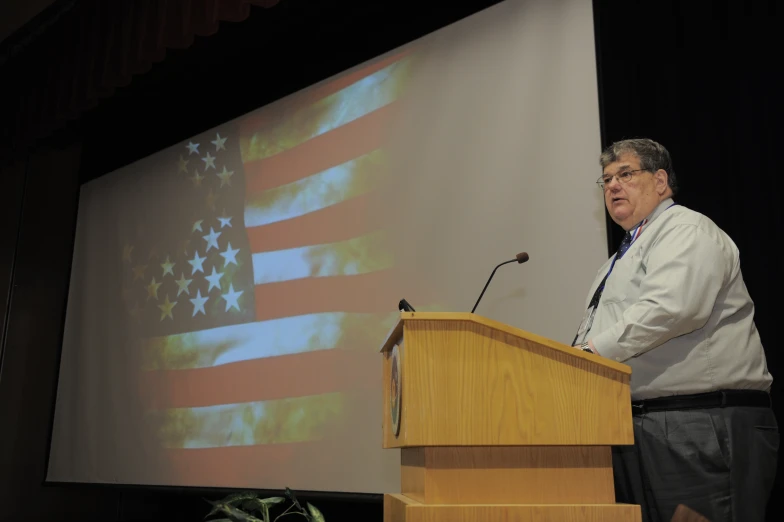 a man speaking at podium with an american flag projected on wall