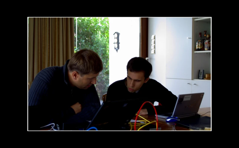 two men sitting at a table looking at a laptop