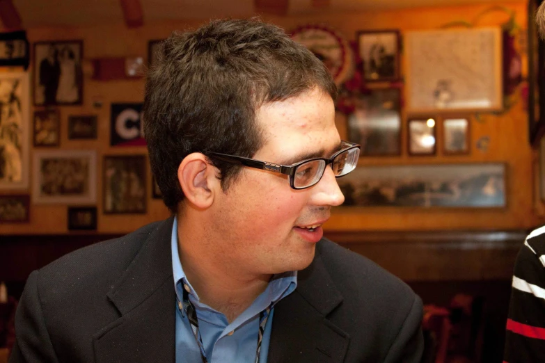 man with short dark hair and glasses, sitting in a booth smiling