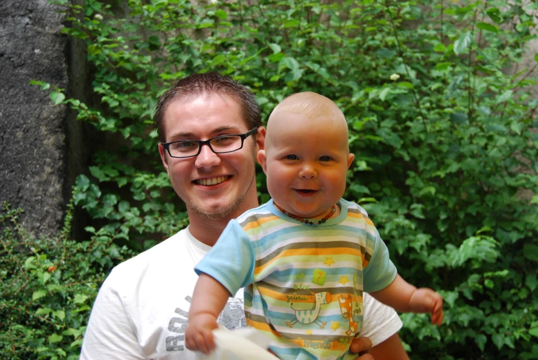a man holding a baby standing next to a lush green forest