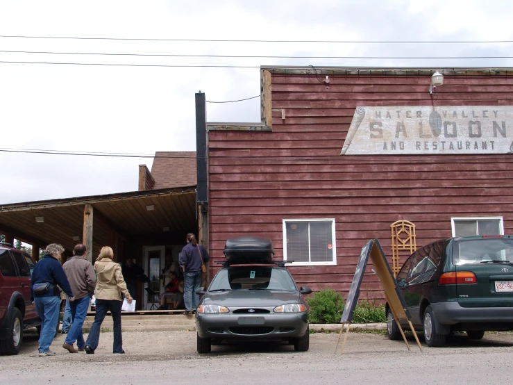 three people stand near two parked cars in front of the saloon