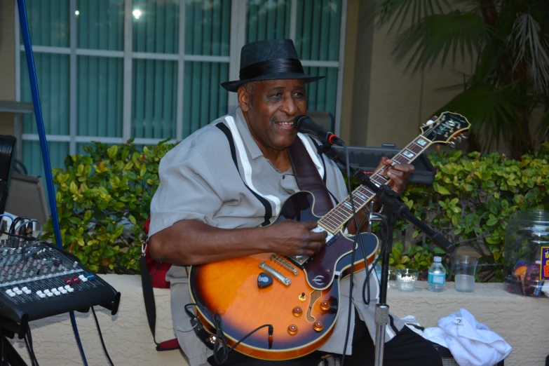 man in fedora with guitar and microphone in outdoor setting