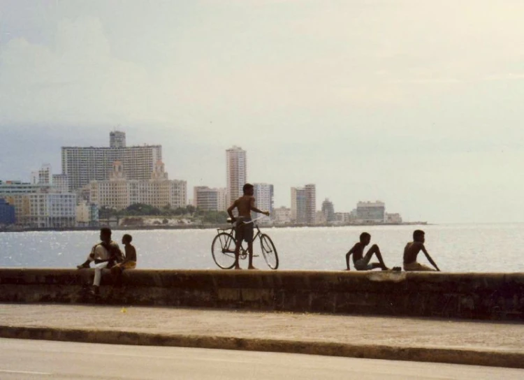 boys sit and stand near a beach while another is riding his bike