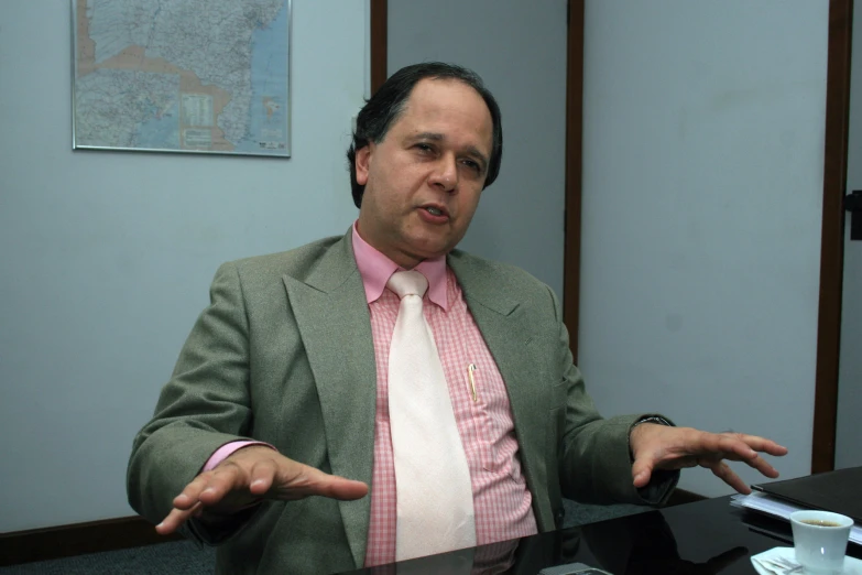 man in grey suit and pink shirt with hands extended, at desk