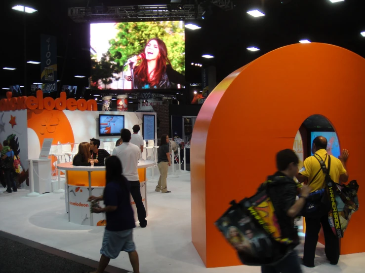 people walking around a trade fair that has a tv screen in the background