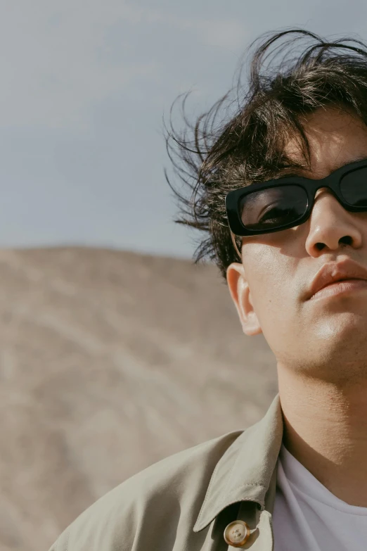 a man wearing sunglasses standing in front of a mountain, woo kim, bad bunny, desert wind, zoomed in