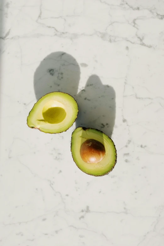 an avocado cut in half on a table, trending on pexels, hyperrealism, smooth marble surfaces, single light, white, olive