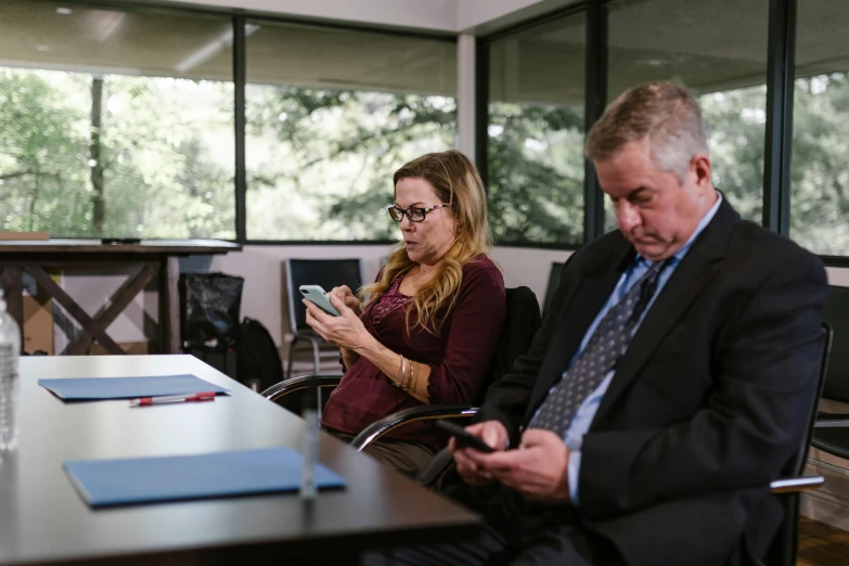 a man and a woman sitting at a table looking at their cell phones, a photo, sitting in dean's office, background image, realistic »