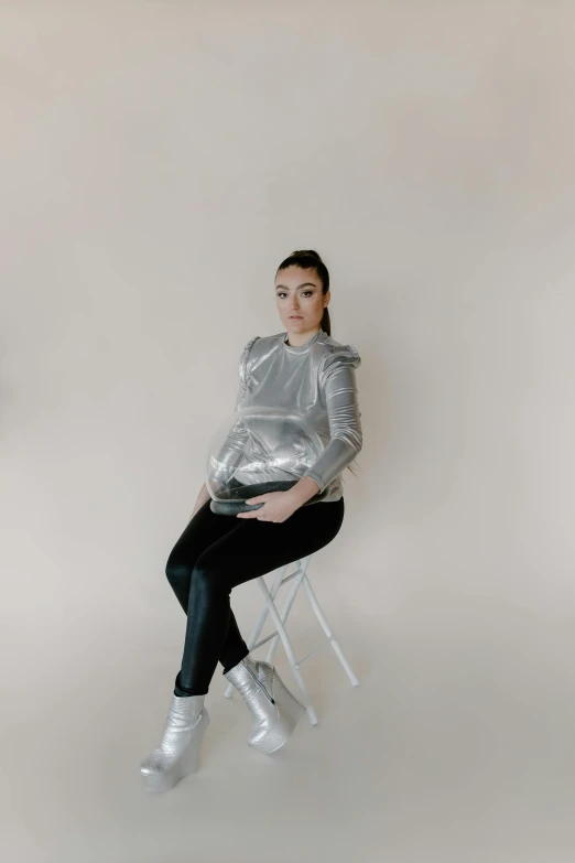 a woman sitting on top of a white chair, inspired by Russell Dongjun Lu, trending on pexels, silver space suit, wearing a designer top, maternal, front facing