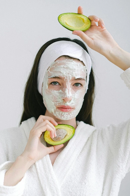 a woman holding a slice of avocado in front of her face, trending on pexels, renaissance, face mask, wearing a white robe, makeup, square facial structure