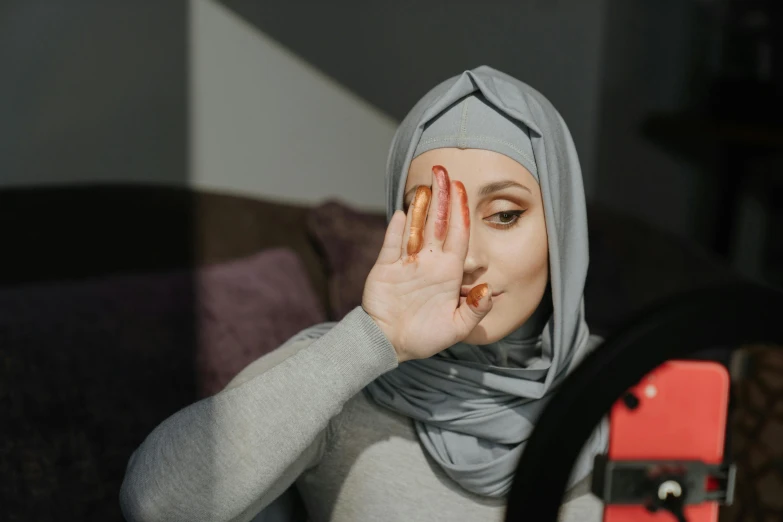 a woman in a hijab covering her face with her hands, a picture, trending on pexels, hurufiyya, putting makeup on, gordon freeman as a woman, triangle makeup, with round face