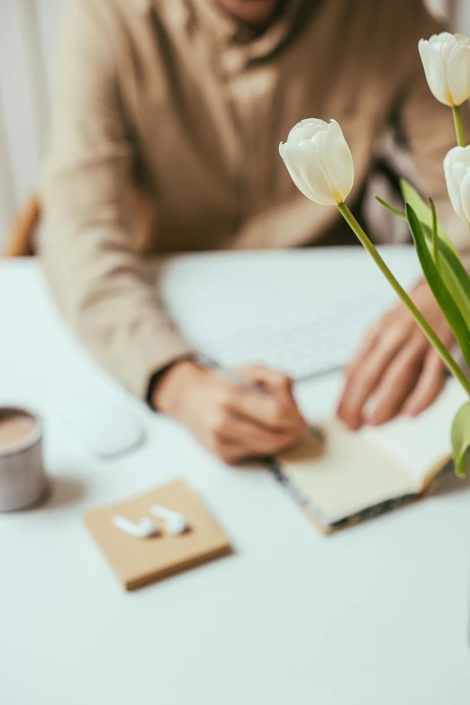 a man sitting at a table writing on a piece of paper, a still life, trending on unsplash, romanticism, tulip, cream and white color scheme, sat in an office, woman in flowers