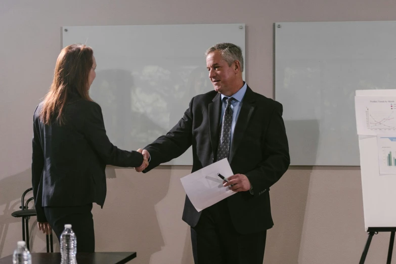 a man and a woman shaking hands in front of a whiteboard, private press, ray swanland, background image, formal attire, awarded