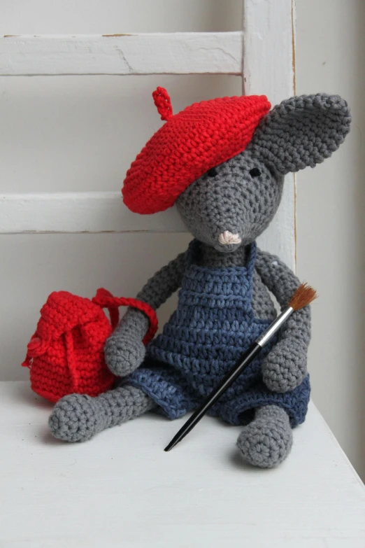 a crocheted mouse is sitting on a chair, a picture, wearing a french beret, paintbrush, school bag, red and blue garments