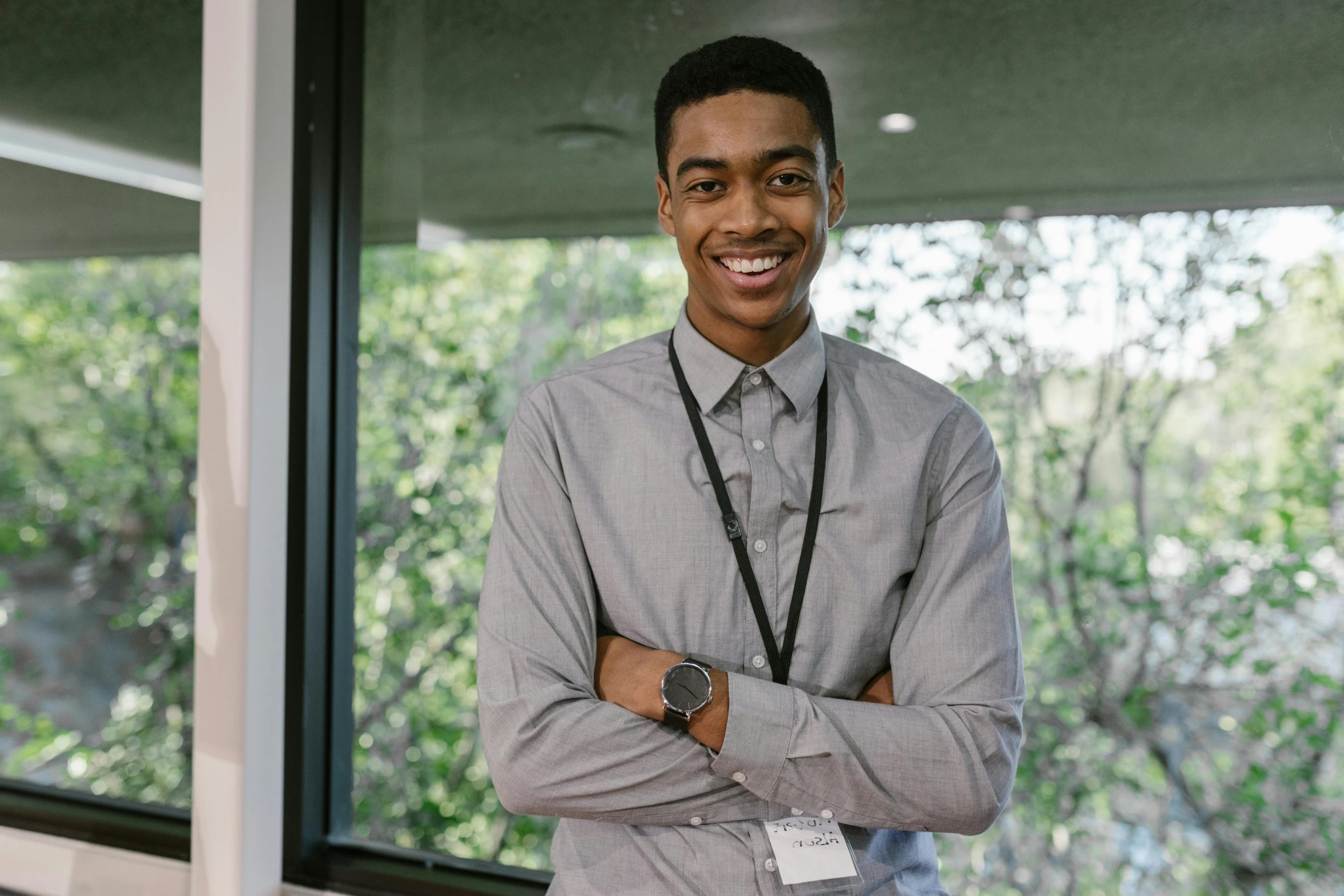 a man standing in front of a window with his arms crossed, a picture, black teenage boy, wearing a shirt with a tie, lachlan bailey, smiling for the camera