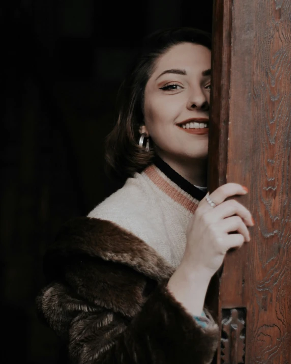 a woman in a fur coat leaning against a door, inspired by Elsa Bleda, trending on pexels, renaissance, half onesided smile, old timey, smiling down from above, androgynous person