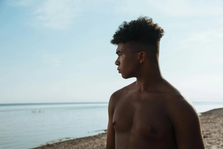 a man standing on a beach next to a body of water, by Carey Morris, pexels contest winner, black teenage boy, with textured hair and skin, pictured from the shoulders up, gay