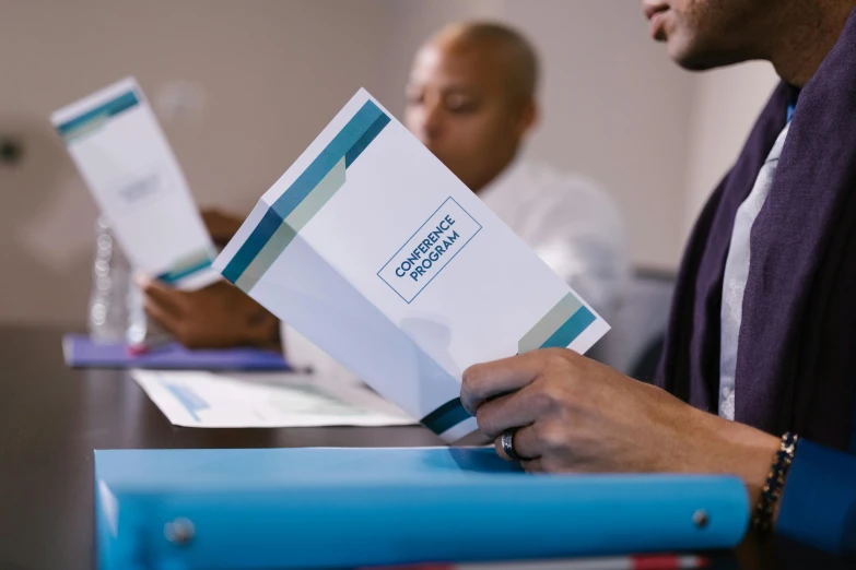 a man holding a folder while sitting at a table, unsplash, private press, teal paper, foreground focus, promotional material, royal commission