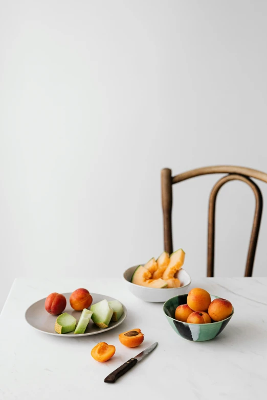 a table topped with bowls of fruit next to a knife, inspired by Constantin Hansen, unsplash, visual art, clean white background, two plastic chair behind a table, peaches, snacks