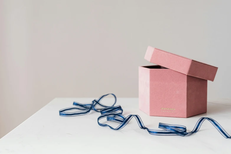 a pink box sitting on top of a white table, by Eden Box, unsplash, private press, blue and yellow ribbons, terracotta, dark. no text, pulling strings