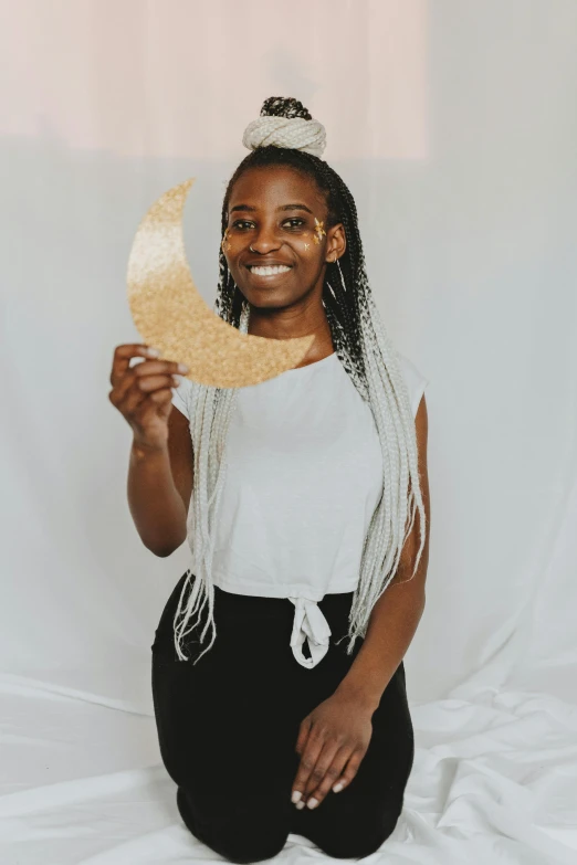 a woman sitting on a bed holding a paper moon, by Dulah Marie Evans, standing in gold foil, headshot profile picture, while smiling for a photograph, with brown skin