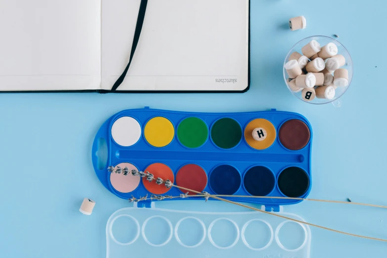 an open notebook sitting on top of a blue table, a child's drawing, inspired by Damien Hirst, trending on pexels, paint tubes, detail on scene, water colour, art set