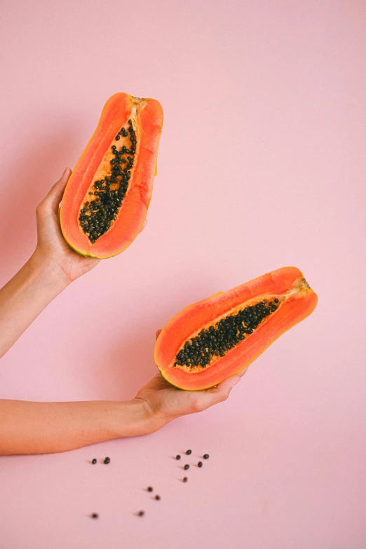 a person holding two slices of papana on a pink surface, by Julia Pishtar, trending on pexels, hurufiyya, orange and black, abundant fruition seeds, tropical, beans