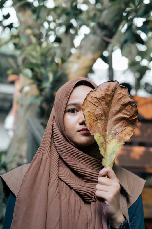 a woman holding a leaf in front of her face, by Basuki Abdullah, unsplash contest winner, hurufiyya, wearing a brown, model posing, hijab, a handsome