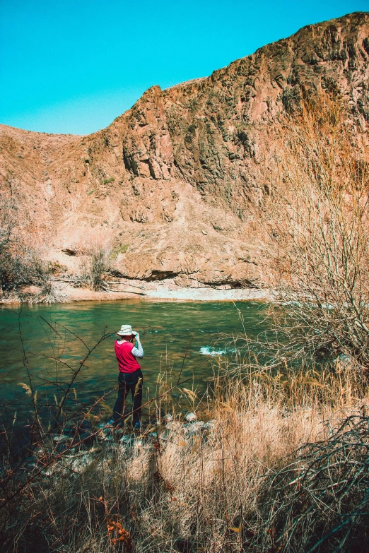 a man standing on a rock next to a river, new mexico with a yellow filter, people angling at the edge, red river, cliffs